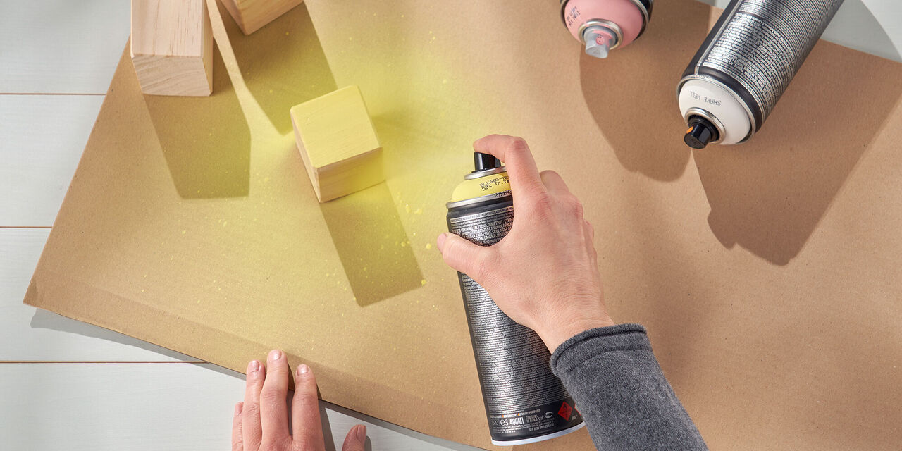 Eine Hand sprüht gelbe Farbe aus einer Spraydose auf braunen Karton, daneben stehen Holzblöcke.