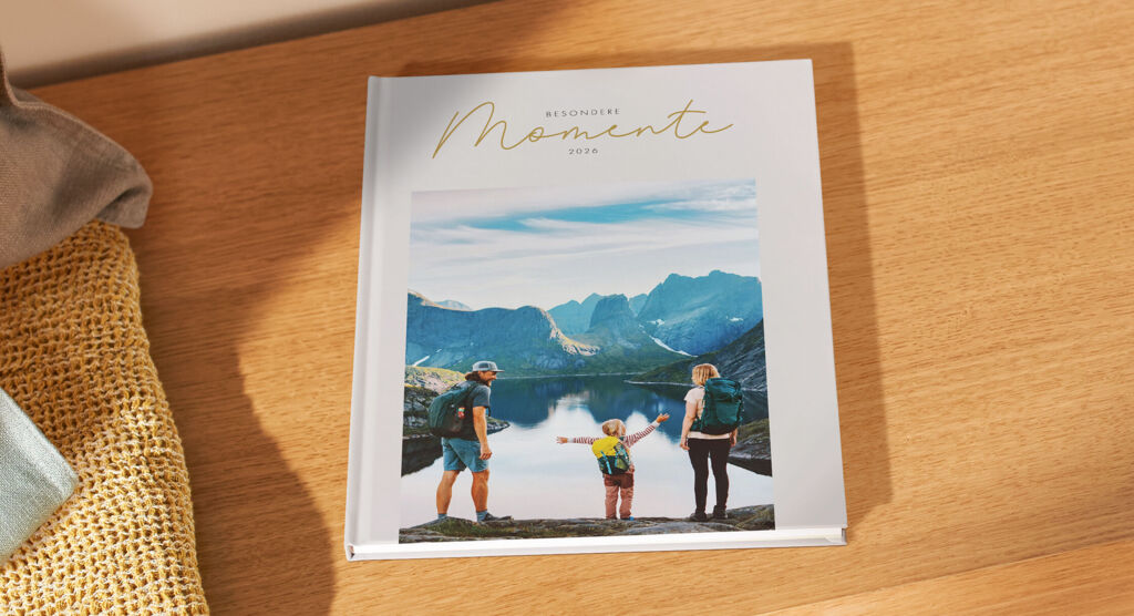 Auf einem Holztisch liegt ein weißes Fotobuch mit der Aufschrift „Besondere Momente 2026“ und dem Foto einer Familie vor einem Bergsee.