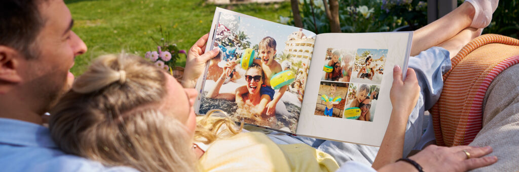 Ein Paar schaut sich gemeinsam im Garten ein Fotobuch mit fröhlichen Sommer- und Schwimmbad-Fotos an.