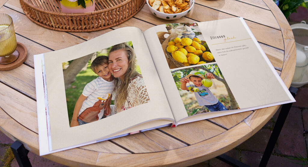 Ein geöffnetes Fotobuch liegt auf einem Holztisch im Garten und zeigt Fotos von einer Frau mit Kind, Zitronen und einem Jungen.