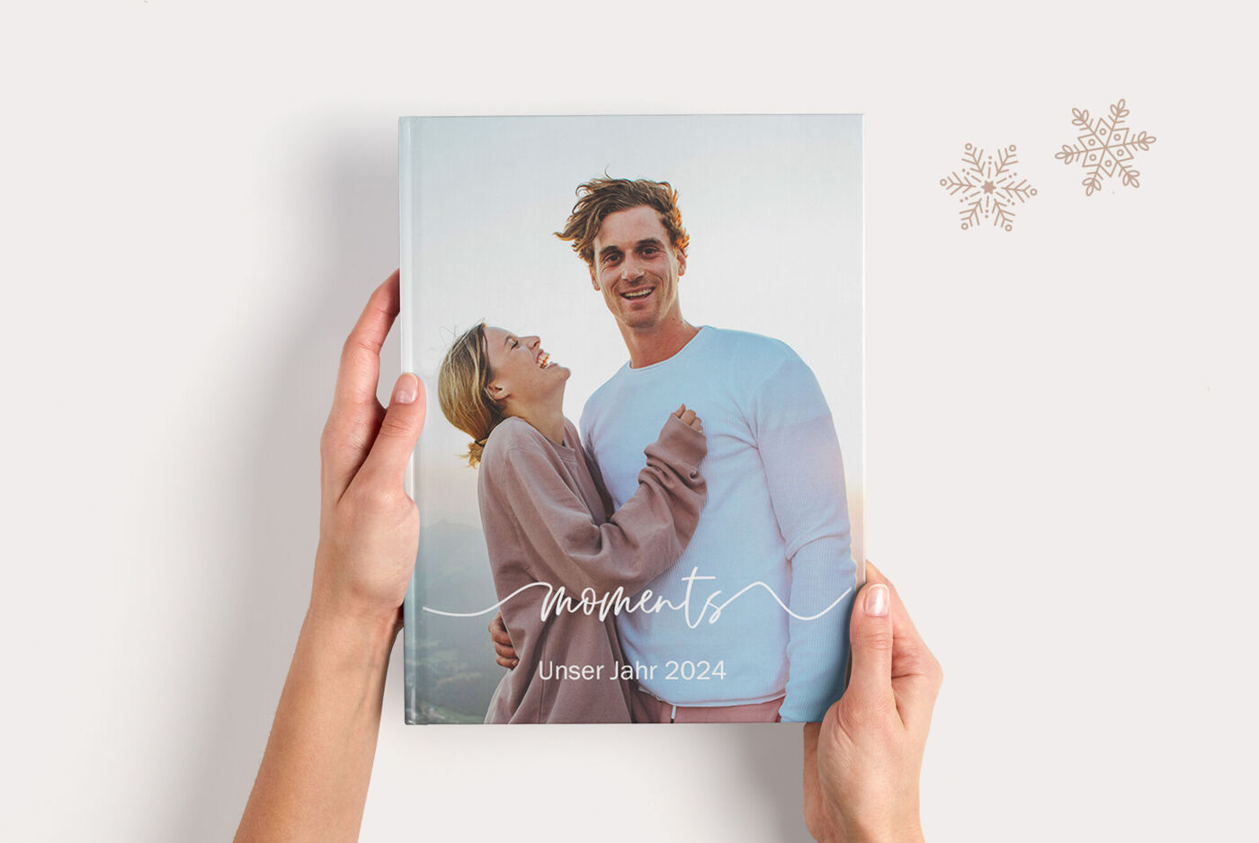 "Hände halten ein CEWE FOTOBUCH mit einem Paarfoto und dem Schriftzug „Moments Unser Jahr 2024“. "