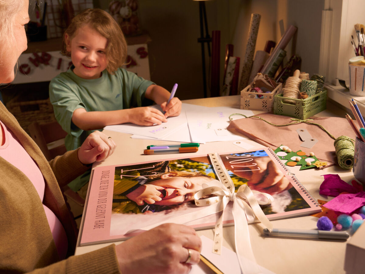 Eine Frau sitzt mit einem Kind an einem Tisch. Das Kind malt, während die Frau einen Fotokalender mit einer Schleife verziert.