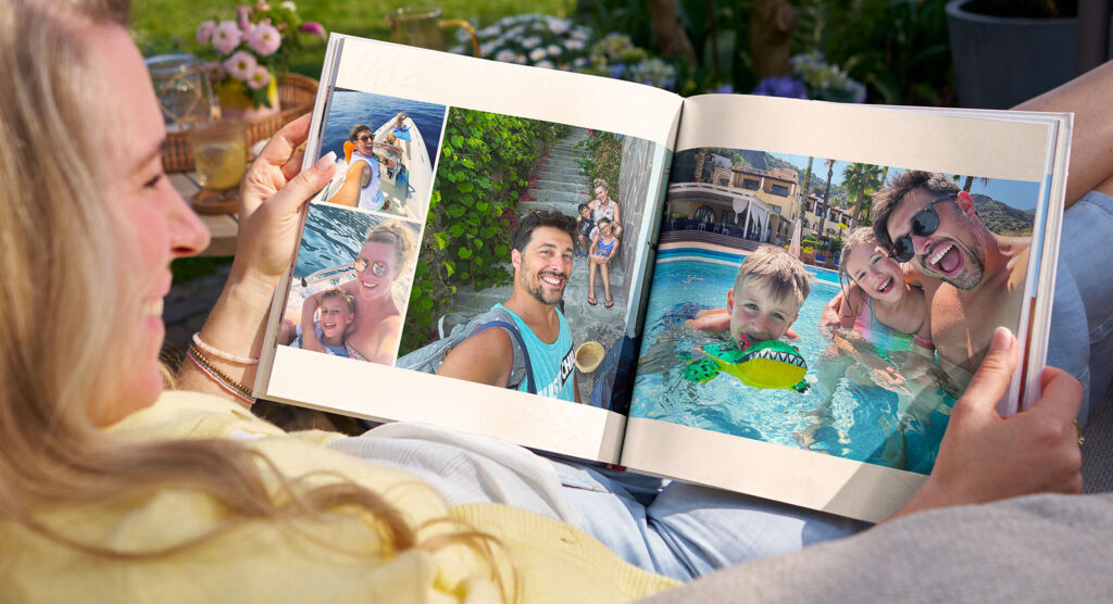 Eine Frau sitzt entspannt im Garten auf einem Sessel und betrachtet ein Fotobuch mit fröhlichen Urlaubsbildern einer Familie am Pool.