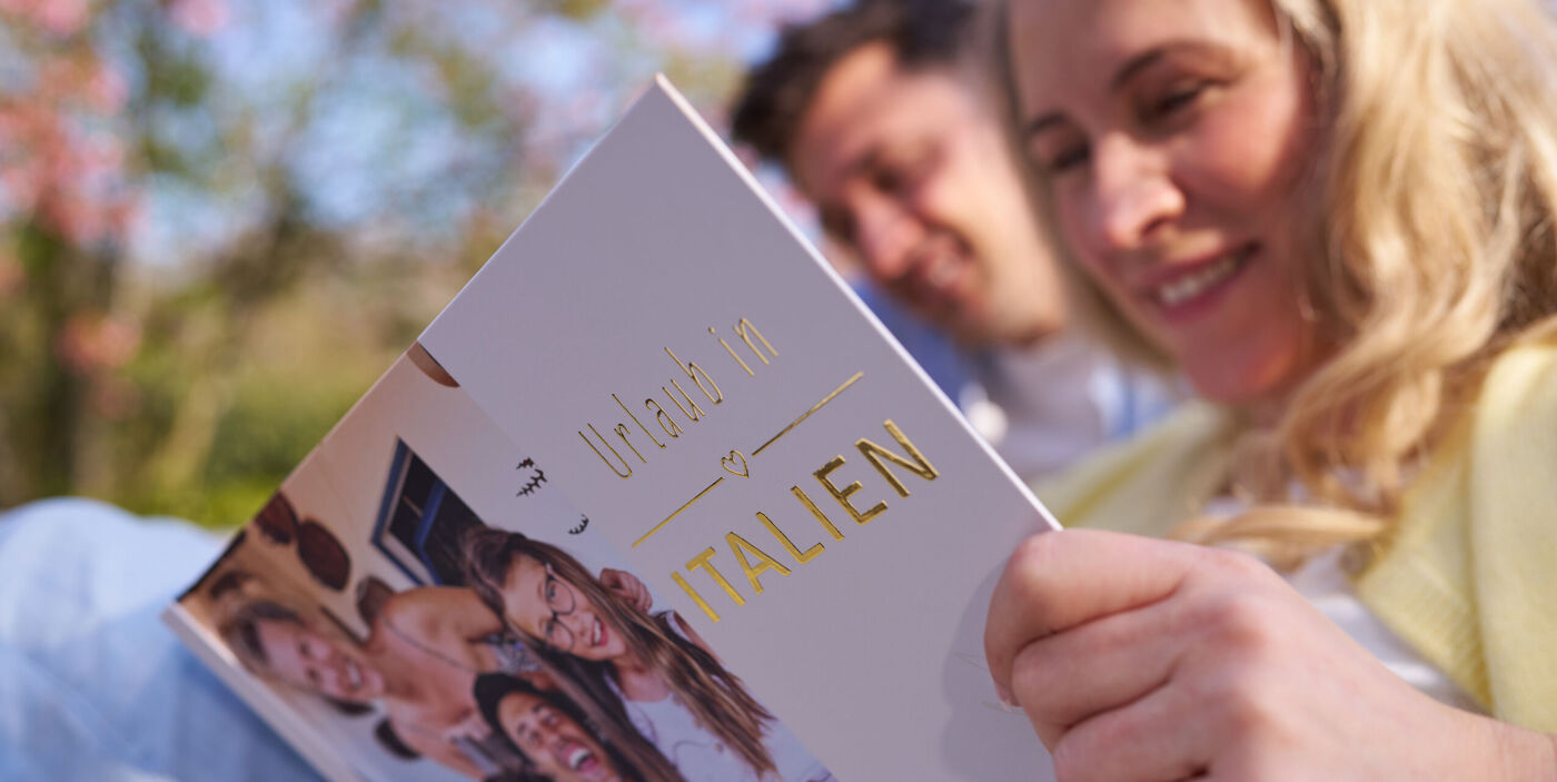 Zwei Personen sitzen im Freien und betrachten gemeinsam ein Fotobuch mit Familienfoto auf dem Cover.